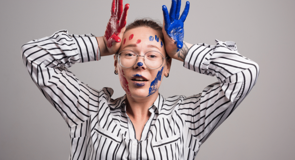 Young woman with glasses and a striped shirt, smiling playfully with colorful red and blue paint on her face and hands, symbolizing the positive side of self-control and self-respect through creative self-expression.
