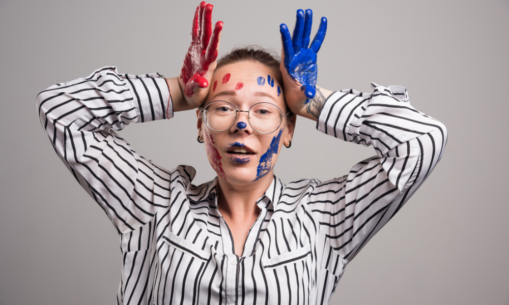 Young woman with glasses and a striped shirt, smiling playfully with colorful red and blue paint on her face and hands, symbolizing the positive side of self-control and self-respect through creative self-expression.